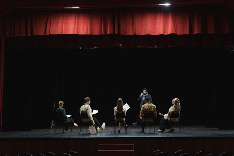 Group of actors on stage reading lines and sitting in a circle.
